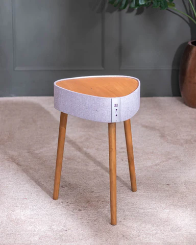 Small round stool with wooden legs and a light gray upholstered seat against a dark gray wall.